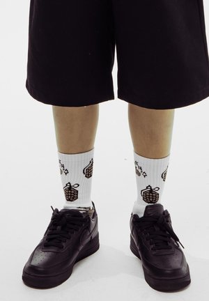 Zapatillas negras de Nike, calcetines blancos con estampado de piñas marrones y pantalones cortos negros hasta la rodilla. Textura suave y diseño casual.