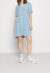 Abito azzurro chiaro a maniche corte con motivo floreale, scollo a V e vita arricciata. Abbinato a sneaker bianche alte.