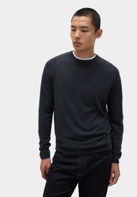 Dunkelblaues Rundhals-Pullover aus weichem Stoff mit glatter Textur. Figurbetonter Schnitt mit langen Ärmeln, über einem weißen T-Shirt getragen.