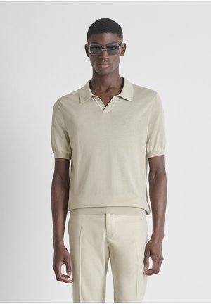 Uomo che indossa una polo beige a maniche corte e pantaloni abbinati, con occhiali da sole scuri, in piedi contro uno sfondo chiaro e uniforme.