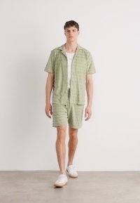 Lichtgroen shirt met korte mouwen en bijpassende korte broek met gestructureerde strepen; gedragen met een wit onderhemd en witte sneakers. Eenvoudig, casual ontwerp.