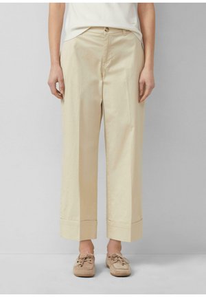 Personne portant un pantalon beige à jambes larges avec revers et des mocassins beiges à lacets, debout devant un fond gris clair.