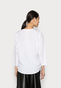 Witte blouse met driekwart mouwen, ronde halslijn en een subtiele plooival langs de achterkant. Draagbaar over een zwarte leren rok.
