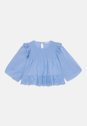 Lyseblå bluse med flagrende ærmer, smocked forstykke og lagdelt kant med hulmønster. Nøglerafslutning bagpå.