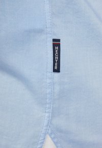 Lichtblauwe shirt stof met een gladde textuur. Heeft een gestikte naad en een klein merketiket met "HECHTER" in het wit op een donkerblauwe achtergrond.