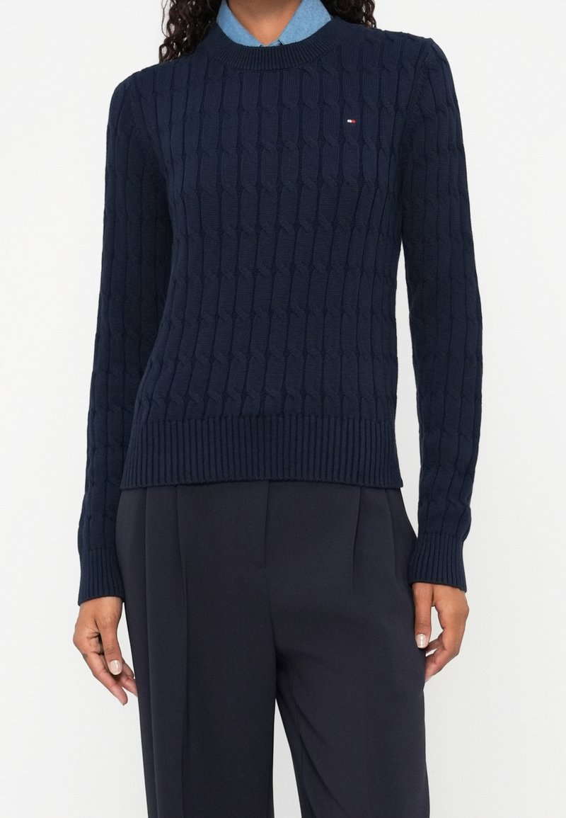 Femme portant un pull en maille torsadée bleu marine sur une chemise à col bleu clair, assorti à un pantalon sur mesure bleu marine foncé.