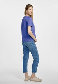 Fioletowa koszulka z krótkim rękawem, luźny krój, zestawiona z jasnoniebieskimi dżinsami w prostym kroju, podwiniętymi na kostkach, uzupełniona beżowymi sportowymi butami.