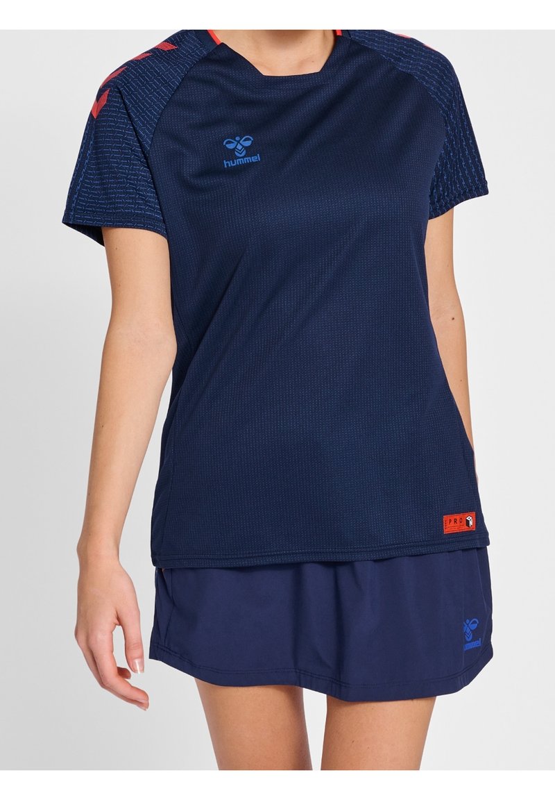Femme portant un maillot de sport bleu marine avec des accents rouges et une jupe de sport bleu marine assortie sur un fond uni.