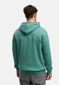 Tyrkis hettegenser med teksturert stoff, ribbestrikkede ermer og nederkant. Har en glatt overdel og en avslappet passform.