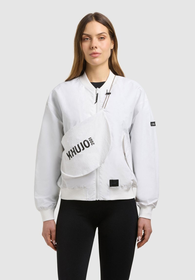 Femme portant une veste blanche zippée et un sac bandoulière blanc avec texte noir, associée à un pantalon noir, sur un fond gris uni.