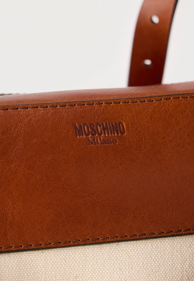 Κοντινό πλάνο του λουριού καφέ δερμάτινης τσάντας με ανάγλυφο το "MOSCHINO Milano" πάνω από μπεζ υφαντή υφασμάτινη επιφάνεια.