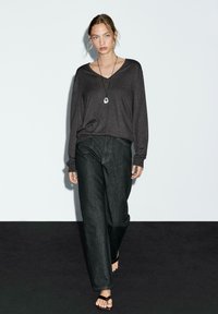 Maglia grigia scuro a maniche lunghe con scollatura a V, abbinata a jeans neri in denim larghi. Il modello indossa sandali bassi e una collana con pendente lungo.