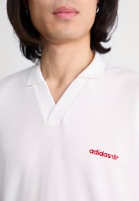 Bílá polokošile s designem ve výstřihu do V, s červeným vyšitým logem "adidas" na levé části hrudi. Hladká struktura látky.