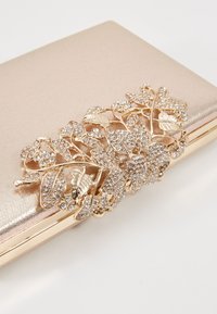 Pochette dorée avec une décoration florale et feuillage scintillant recouverte de petits cristaux transparents sur fond blanc.