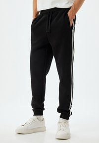 Pantalones de chándal negros hechos de tejido suave, con rayas laterales blancas, cinturilla elástica con cordón y puños acanalados. Combinados con zapatillas blancas.