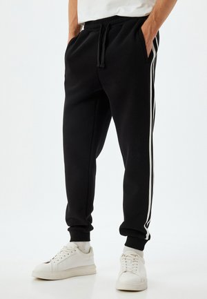 Sorte jogger-bukser lavet af blødt stof, med hvide sidelister, elastisk talje med snøre og ribbede mansjetter. Båret med hvide sneakers.