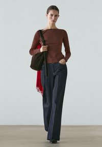 Kvinde der går iført en tætsiddende brun langærmet top, mørke vidde ben jeans, sorte sko og bærer en brun skuldertaske med et rødt tørklæde.