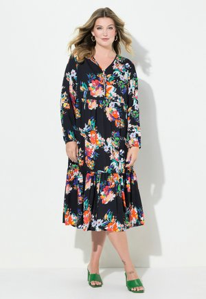 Florales Kleid in Schwarz mit lebhaften Farben; lange Ärmel, V-Ausschnitt, Knopfdetails und gestuftem Rock. Kombiniert mit grünen Absatzsandalen.