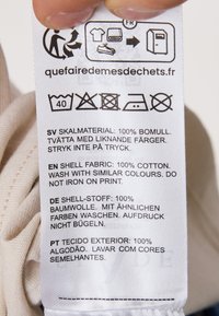 Vaskeanvisning på bomuldslabel med flersprogede instruktioner, der indeholder symboler for vedligeholdelse; farverne er primært hvide og beige.
