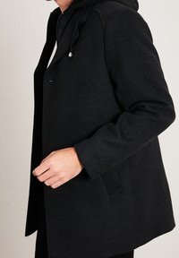 Cappotto in lana nera con cappuccio, caratterizzato da una vestibilità comoda, tasche laterali e sottili dettagli di cucitura sulle spalle. La texture appare morbida.