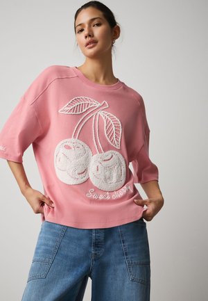 Jeune femme portant une chemise oversize rose avec des cerises blanches brodées et le texte « Sweet cherry », associée à un jean bleu ample.