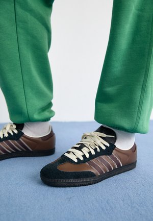 Persoon die bruine en zwarte sneakers draagt met crèmekleurige veters, witte sokken en een groene broek, staande op een blauw tapijtvloer.