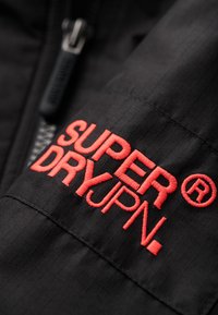 Chaqueta de tela negra con un acabado texturizado, que presenta el logotipo bordado en rojo "SUPER DRYJPN" y un detalle de cremallera.