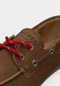 Καφέ καστόρινο παπούτσι του σκάφους με κόκκινες και λευκές κορδόνια περασμένα μέσα από ασημένιες μεταλλικές τρύπες σε απλό φόντο.