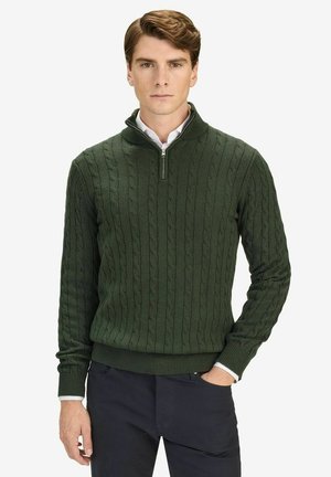 Mand iført en mørkegrøn kabelstrikket pullover med kvart-lynlås over en hvid skjorte, kombineret med mørke bukser, stående mod en ensfarvet baggrund.