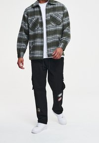 Camicia di flanella a quadri verdi con due tasche sul petto, indossata su una maglietta bianca, abbinata a pantaloni cargo neri con toppe ricamate e sneaker bianche.
