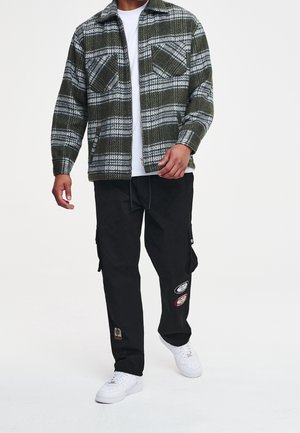 Camisa de franela a cuadros verdes con dos bolsillos en el pecho, llevada sobre una camiseta blanca, combinada con pantalones cargo negros con parches bordados y zapatillas blancas.