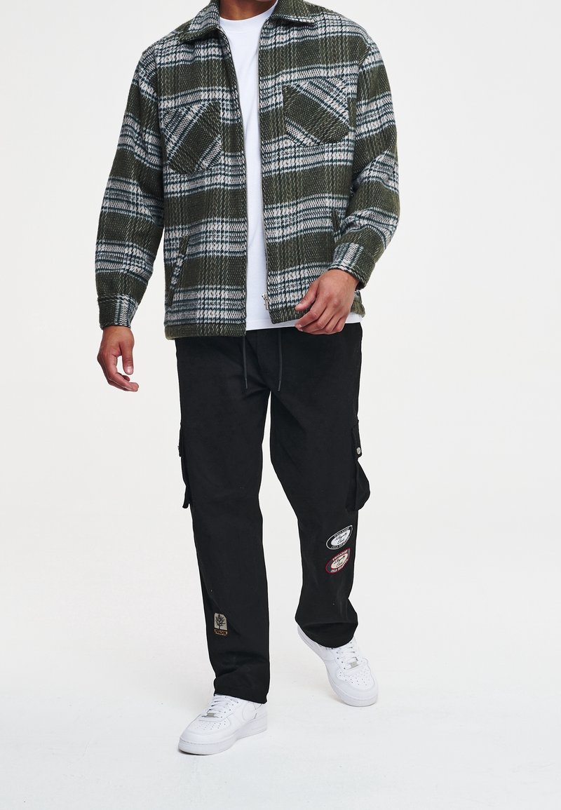 Camicia di flanella a quadri verdi con due tasche sul petto, indossata su una maglietta bianca, abbinata a pantaloni cargo neri con toppe ricamate e sneaker bianche.