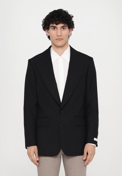 Schwarzer taillierter Blazer mit spitzen Revers, zwei Knöpfen und Seitentaschen. Getragen über einem weißen Hemd, kombiniert mit hellbeigen Hosen. Glatter Stoff.