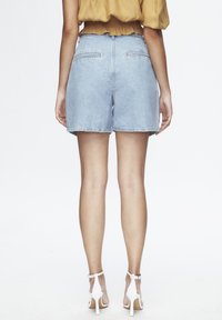 Shorts vaqueros de tiro alto en azul claro, con bajo deshilachado, dos bolsillos traseros y un ajuste holgado, combinados con tacones blancos con tiras.