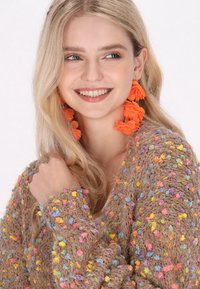 Brun texturerad cardigan med flerfärgade pom-poms; modell som bär klara orangea blommörhängen, håret nedsläppt, ler.
