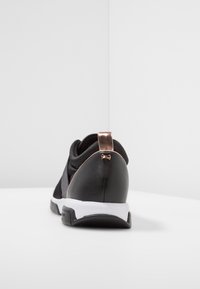 Baskets noires avec une tige en tissu texturé noir, un talon lisse noir, et un accent en or rose sur la languette du talon. Semelle blanche.