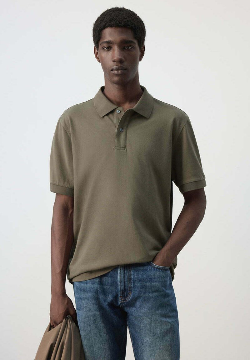 Jeune homme portant un polo vert olive et un jean bleu, tenant une veste assortie dans une main, sur un fond clair uni.