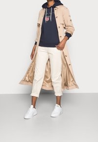 Trench-coat beige avec boutons, sweat à capuche bleu marine avec logo graphique, pantalon crème et baskets blanches. Textures lisses et design en couches.