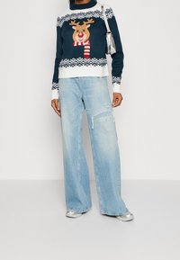 Pletený svetr v tmavě modré barvě s motivem soba, bílými akcenty a geometrickým vzorem. Spárovaný se světle modrými kalhotami s širokými nohavicemi a stříbrnými botami.