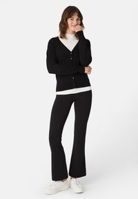 Černý cardigan s výstřihem do V a zapínáním na knoflíky, vrstvený přes bílý top. Doplněno černými zvonovými kalhotami a bílými teniskami.