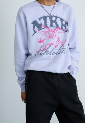 Person iført en lyse lilla sweatshirt med en lyserød vinget hest og tekst "NIKE athletic" kombineret med sorte bukser og lagdelte sølvsmykker.