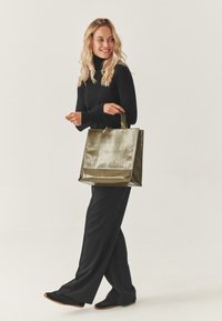 Tragetasche in metallischem Olivgrün mit zwei kurzen Griffen, quadratischer Form, glänzender Textur und einfarbig. Modell trägt schwarze Kleidung.