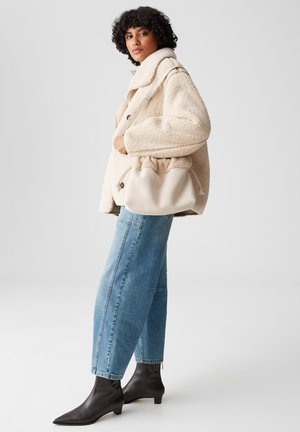 Woman wearing a beige teddy coat, blue wide-leg jeans, black ankle boots, and carrying a beige drawstring shoulder bag.