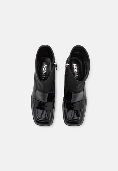 Bottines en cuir verni noir avec un bout carré, présentant un design épuré, une texture lisse et une sangle décorative à l'avant.