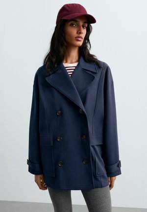 Femme portant un manteau croisé bleu marine, une casquette grenat, une chemise rayée et des leggings gris, debout devant un fond clair uni.