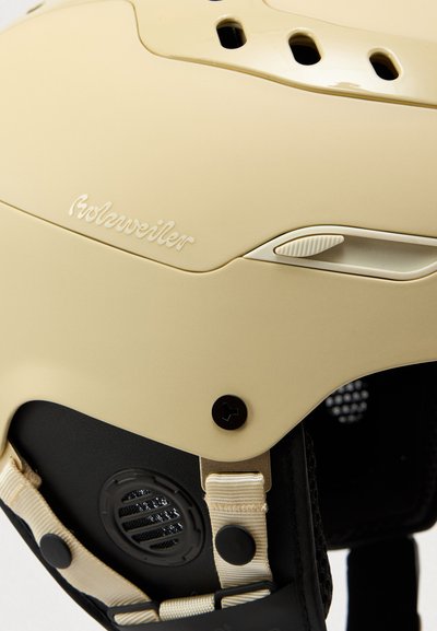 Casco beige con fori di ventilazione, superficie strutturata e cinturino regolabile. Presenta una base nera con una manopola e una ventola simile a un altoparlante.
