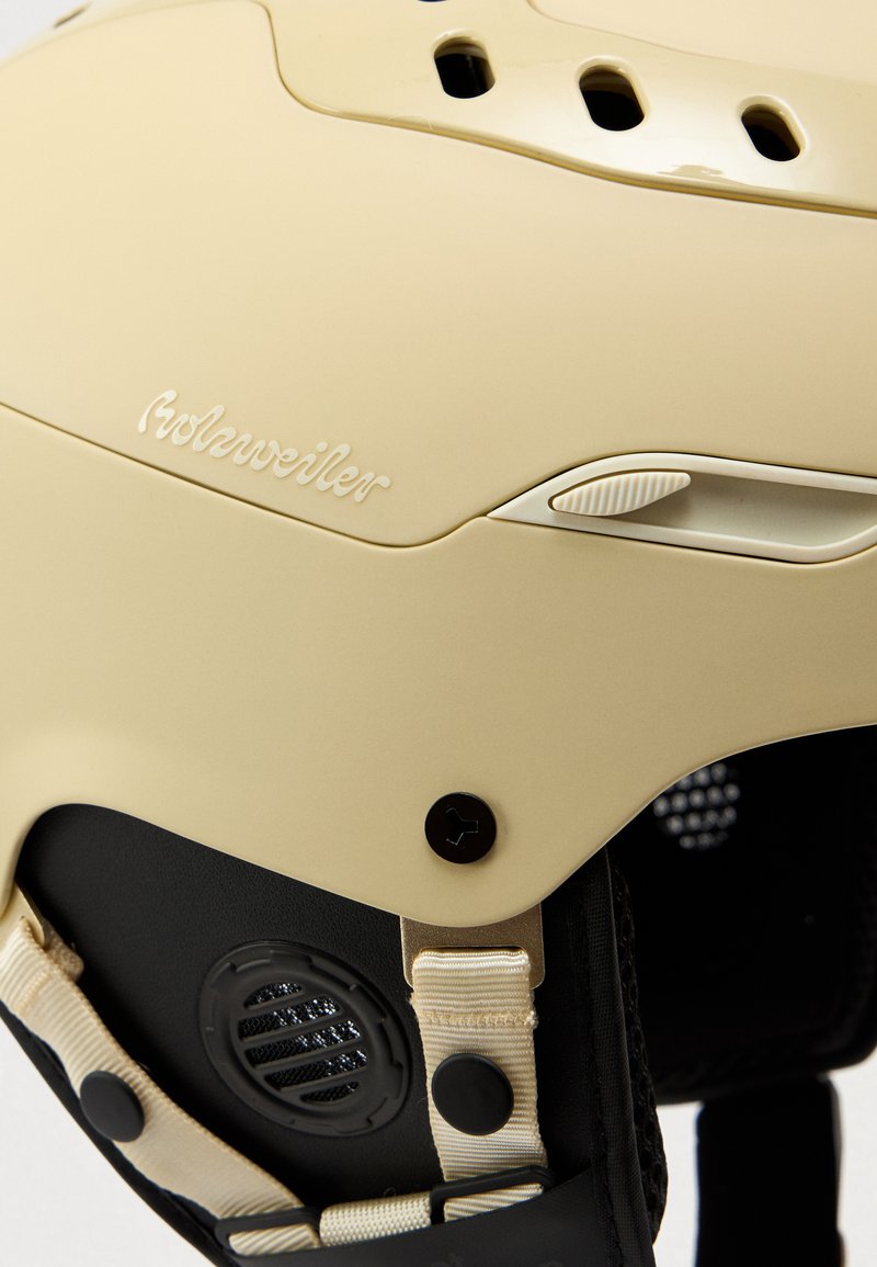 Beige helmet with ventilation holes, textured surface, and adjustable strap. Features a black base with a knob and speaker-like vent.