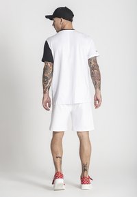 Biela tričko s čiernymi rukávmi, nosené s bielymi šortkami a červenými vzorovanými topánkami. Na ramenách sú tetovania. Jednoduché pozadie.