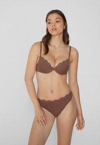 Eine Frau steht in einem passenden Set aus braunem, mit Spitze verziertem BH und Slip, leicht zur Seite schauend vor einem schlichten hellen Hintergrund.