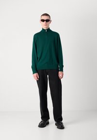 Maglione verde con zip realizzato in tessuto morbido, abbinato a pantaloni neri e scarpe nere. Il modello indossa occhiali da sole scuri e si trova in un ambiente neutro.